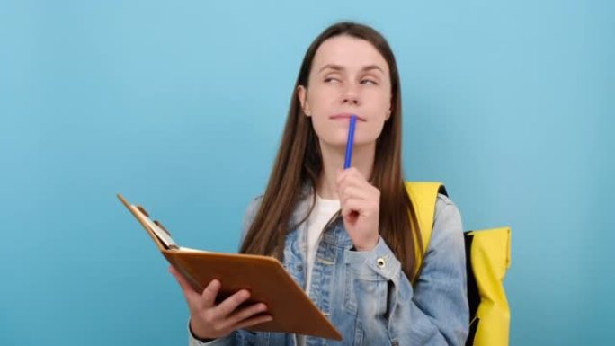 沉思女孩的肖像青少年学生穿着牛仔夹克和黄色背包在练习册笔记中写下，孤立在工作室的蓝色背景墙上。大学学