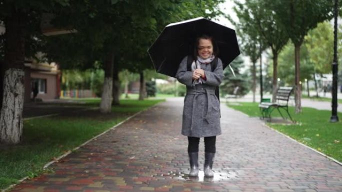 快乐的小人站在城市小巷的雨伞上抬头看着相机，微笑着踩着橡胶靴的脚。白种人小人物在雨天摆姿势的宽镜头肖