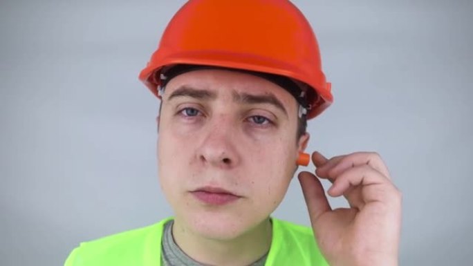 一名戴着安全帽和绿色背心的工人戴上耳塞。工业安全。工业噪声保护。在大声工作中遵守行为规则。橙色耳塞