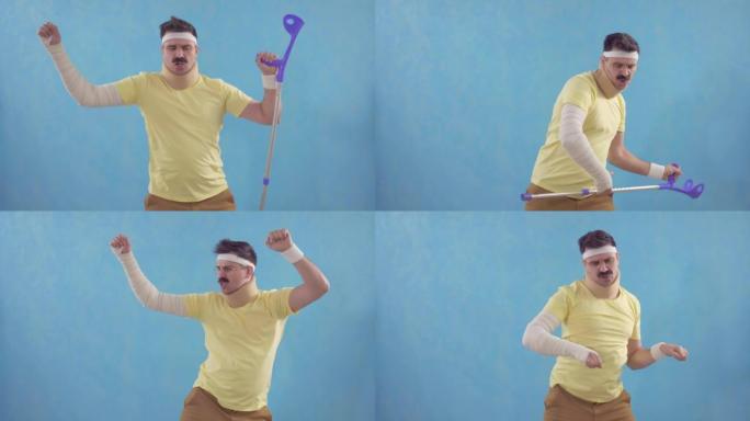 有趣的大胡子男人在运动受伤后，拄着拐杖，用绷带包扎的手站在蓝色背景上在镜头前跳舞