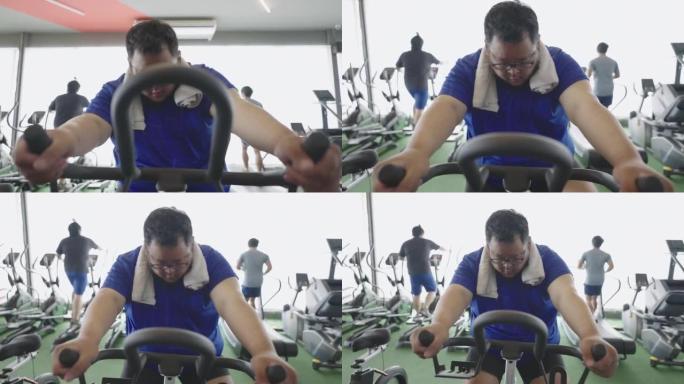超重男子在健身中心骑固定自行车锻炼的前视图。
