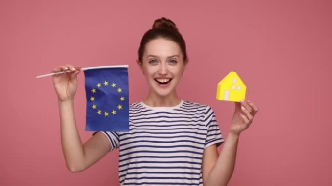 兴高采烈的女子手拿欧盟旗帜和纸房子，梦想着在欧洲买个住处。