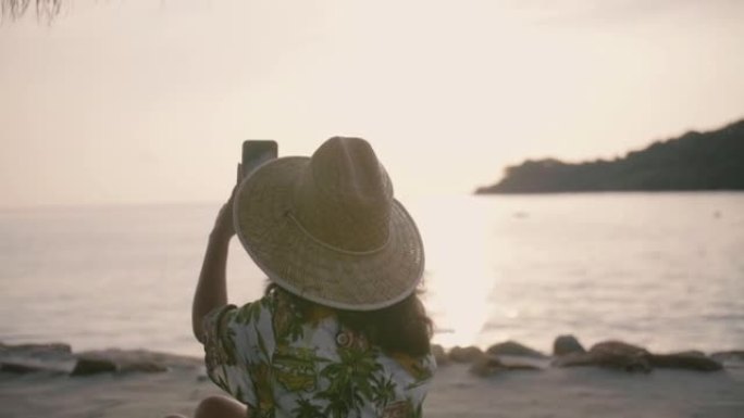 亚洲年轻旅游女子坐在海滩附近，用智能手机拍照。