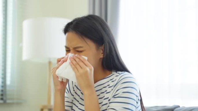 亚洲美女在客厅里流鼻涕和打喷嚏。迷人的年轻女性坐在沙发上感觉不好，过敏，然后在打喷嚏的时候把纸巾盖住