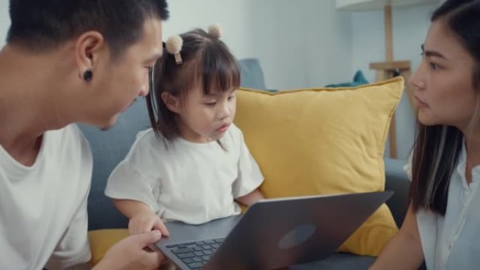 年轻的父母亚洲家庭坐在沙发上享受黄金时光快乐的时刻，和他们的孩子一起在家里的客厅用笔记本电脑看卡通电