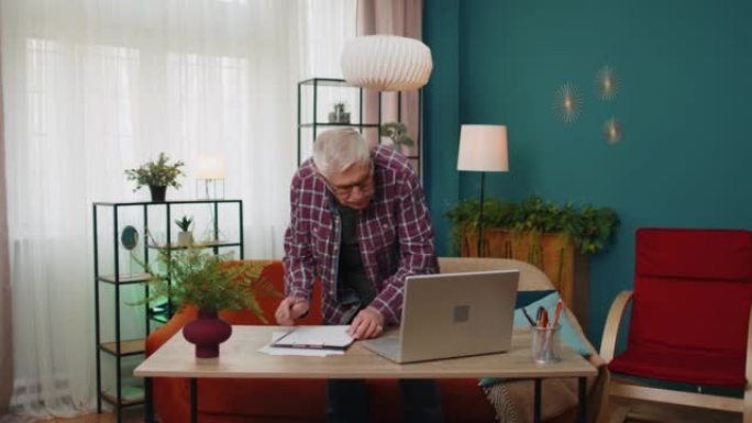 在笔记本电脑上工作的高级祖父在纸上做笔记计算账单