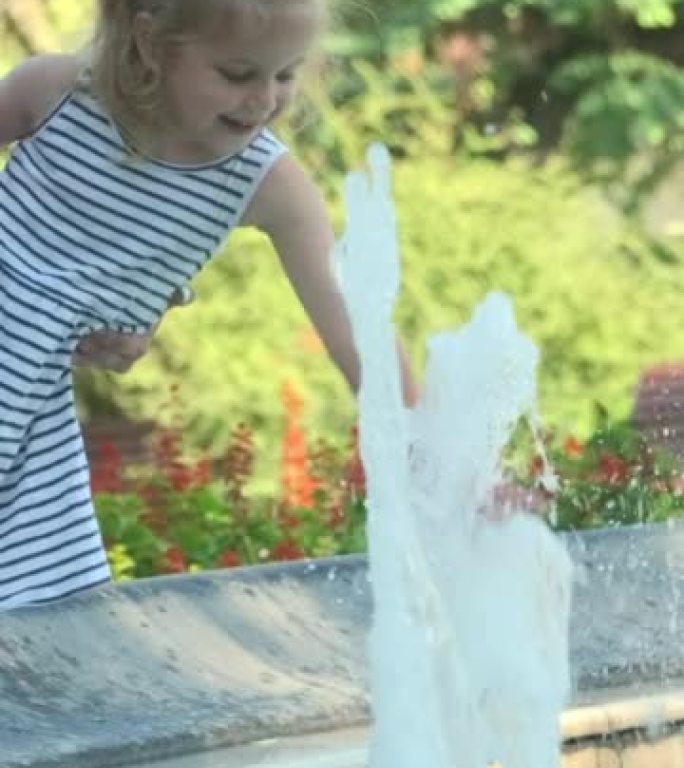 垂直视频: 妈妈的双手抱着微笑的小女儿在喷泉中嬉水。妈妈和小女孩在公园的喷泉里玩耍，微笑着。慢动作。