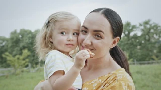快乐的母亲拥抱小女儿，在花园里郁郁葱葱的树上站着吃美味的面包店。女人喜欢在农村与孩子在一起的时间特写