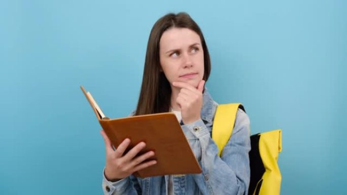 肖像女孩青少年学生穿着牛仔夹克和黄色背包看书思考梦想，在工作室的蓝色背景墙上孤立地摆姿势。高中教育大