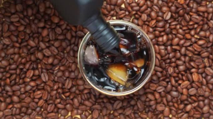 4k视频，在杯子里倒黑咖啡。冷冲泡咖啡的俯视图将咖啡倒入带有咖啡豆背景的冰块上的玻璃杯中。带泡沫的咖