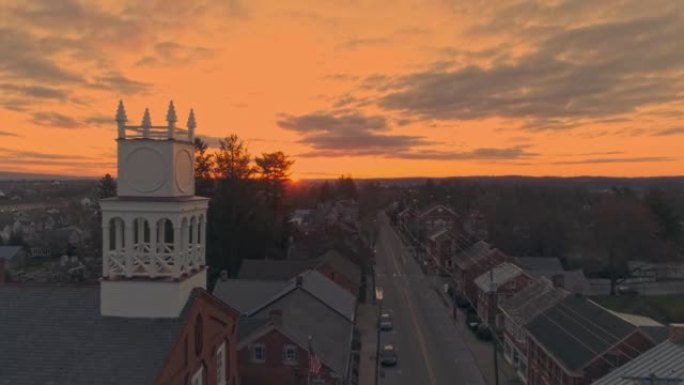 无人机在日出时向后拉一个小镇和一个尖顶的景色，因为它准备打破地平线，春天的日出时橙色和红色