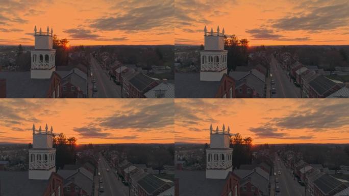 无人机在日出时向后拉一个小镇和一个尖顶的景色，因为它准备打破地平线，春天的日出时橙色和红色