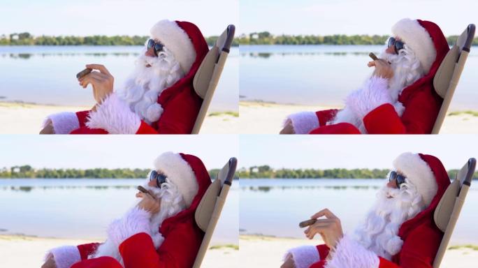 戴着墨镜的圣诞老人躺在湖边海滩的日光浴躺椅上抽雪茄