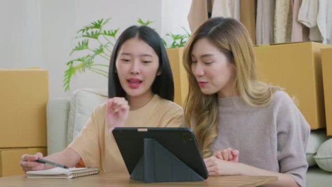 年轻的亚洲夫妇妇女在出售衣服后占收入和支出。几个女人坐在一张小桌子旁，用平板电脑在家做账。