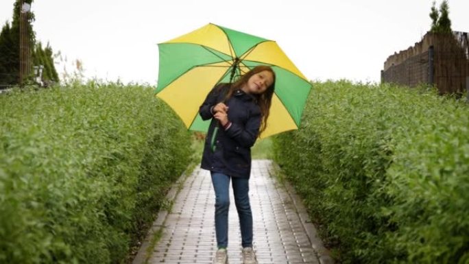 可爱的小女孩在春雨中撑着伞走在绿色的灌木丛中，脸上洋溢着积极的情绪，对着镜头微笑着
