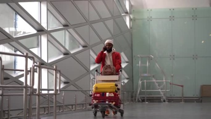 微笑的黑人男子推着行李手推车到达机场后走路，用手机聊天。