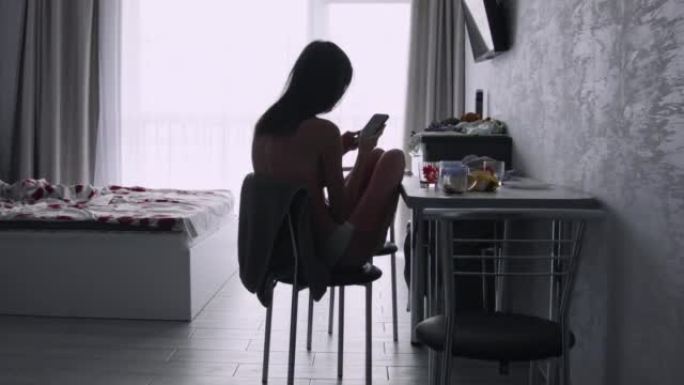 年轻的露背女人坐在卧室的椅子上使用智能手机。