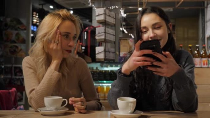 咖啡馆里的两个年轻美女使用手表智能手机聊天社交媒体，而另一个女朋友无聊喝茶
