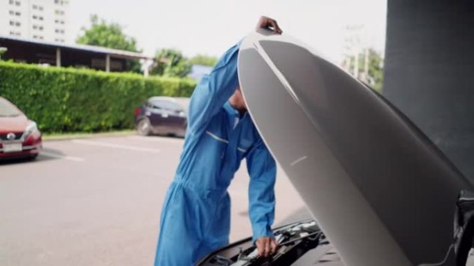 亚洲中年汽车修理工穿着蓝色机械连身衣打开汽车发动机罩检查汽车电池问题，家庭服务