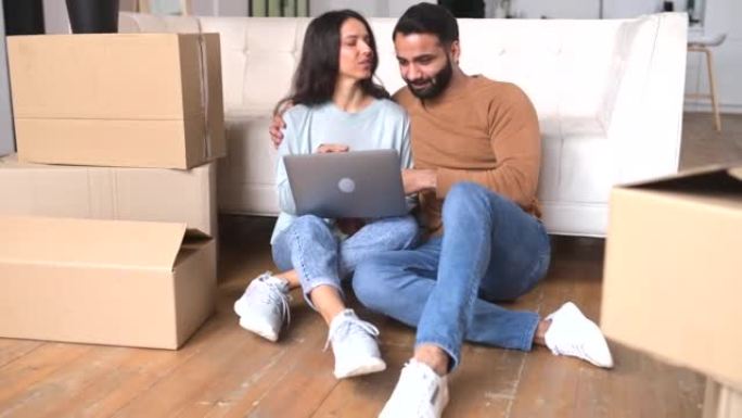 20多岁的印度年轻夫妇一起使用笔记本电脑坐在新公寓的地板上，周围都是纸板箱，多种族的男女朋友在网上选