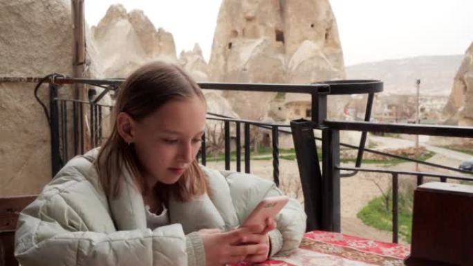 穿着夹克的可爱女孩坐在露天露台咖啡馆餐厅的桌子旁，在智能手机上输入文字。