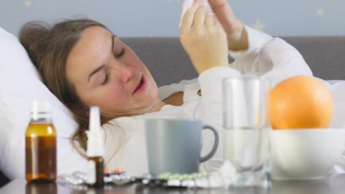 一名患重感冒的年轻女子躺在毯子下的床上，在将鼻涕吹入餐巾纸后使用滴鼻剂。使用鼻子喷雾的女人