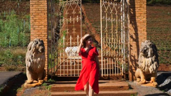 夕阳下，殖民风格的公园里，穿着红色连衣裙的迷人时髦的女人走在带狮子雕塑的锻铁大门附近。时尚理念，欧式