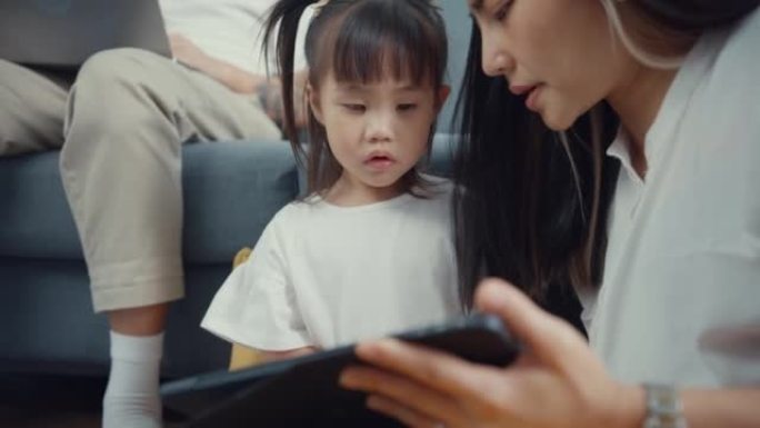 关闭年轻的亚洲父母父亲使用笔记本电脑在家工作母亲使用平板电脑玩游戏，并与女儿一起在家里的客厅看卡通电