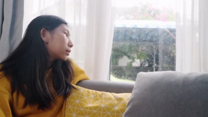 亚洲女孩穿着黄色毛衣坐在沙发上，在雨天透过窗户看，生活方式的概念。