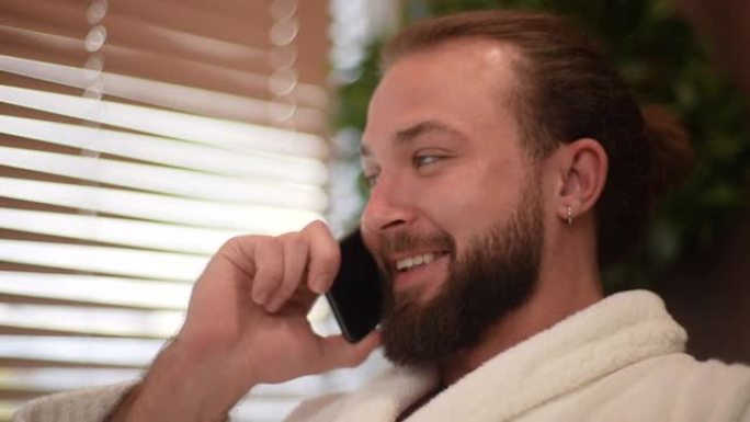 穿着白色浴袍的英俊男性在家里休息时使用移动智能手机交谈的特写脸。微笑的大胡子男人打来电话的特写