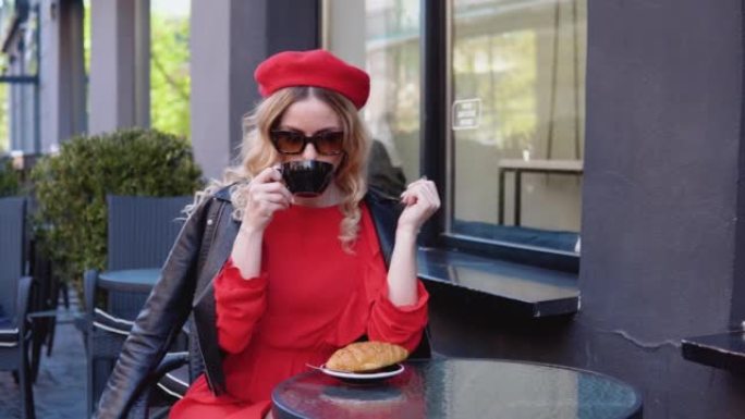 时尚咖啡。金发女郎用羊角面包喝咖啡。餐厅的现代黑色露台。戴着太阳镜和贝雷帽的年轻女子
