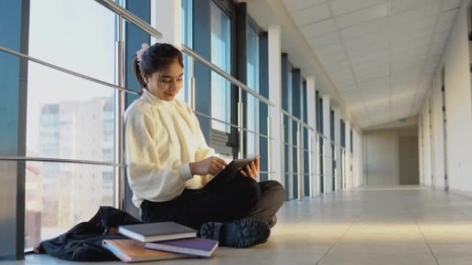 印度女学生坐在地板上，拿着平板电脑在大学或学院。新的现代化功能齐全的教育设施。网络教育的概念