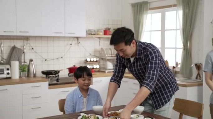 快乐的亚洲人在家里一个现代明亮的餐厅里。当母亲端菜时，父亲接管了盘子，母亲坐下来一起吃饭。