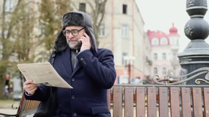 一位老人在城市里享受着寒冷的冬日早晨。一名男子坐在广场的长椅上，看报纸，兴高采烈地用手机与朋友聊天。