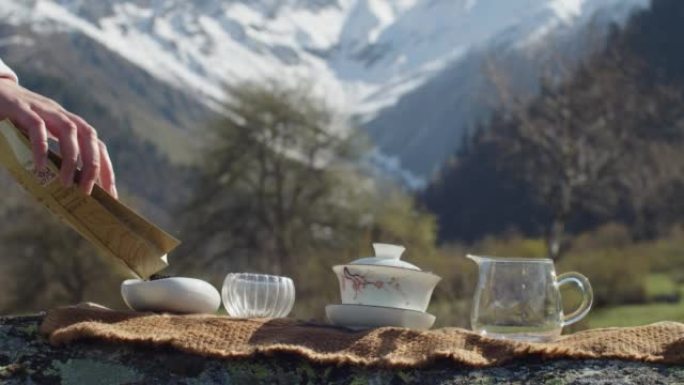 面无表情的女人在雪山的背景下将茶倒入碗中。大自然茶道专用器皿的准备。户外传统中国茶道