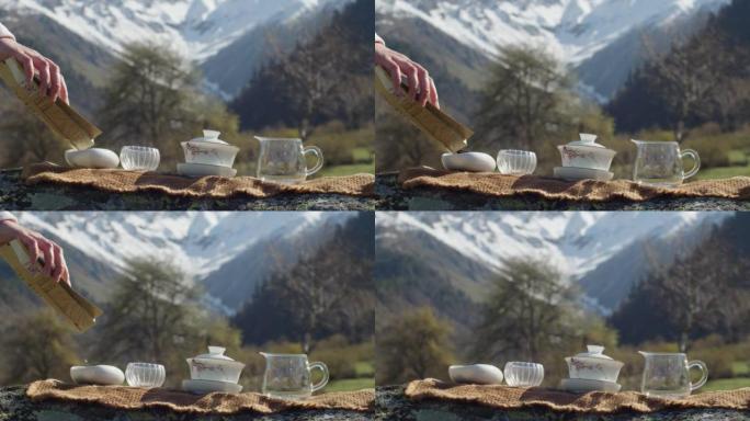 面无表情的女人在雪山的背景下将茶倒入碗中。大自然茶道专用器皿的准备。户外传统中国茶道