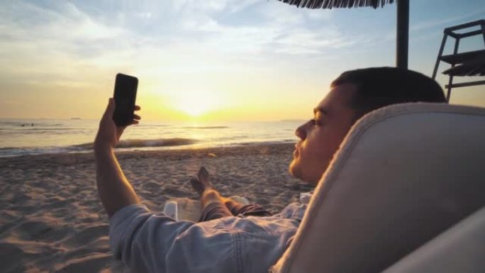 年轻人在日落，日出时在三一热带海滩上的躺椅上享受，并使用智能手机，度假和夏季旅行进行自拍照