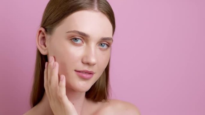 美丽的年轻女子模特用手指触摸她的脸在粉红色的背景。健康清洁护肤理念。