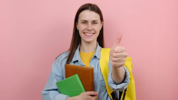 快乐女孩的肖像青少年学生穿着衬衫和黄色背包拿着书显示拇指向上像手势，孤立在工作室的柔和粉色背景上。大