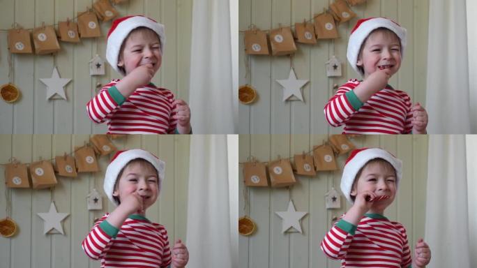 穿着睡衣的男孩，圣诞老人帽子在带有礼物背景的纸质降临日历上吃甜糖果。墙上有手工圣诞日历礼物的小快乐孩