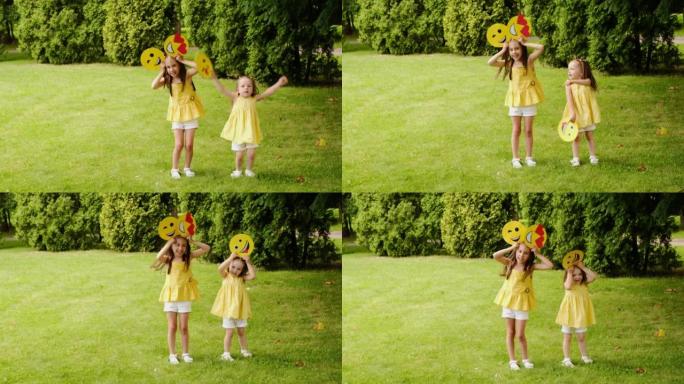 两个有趣的小女孩在公园的草坪上做鬼脸跳舞，手里拿着不同的表情符号