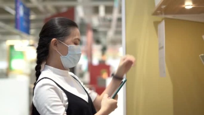 戴着防护口罩的亚洲妇女使用智能手机拍照或扫描QR码以检查百货商店的库存。