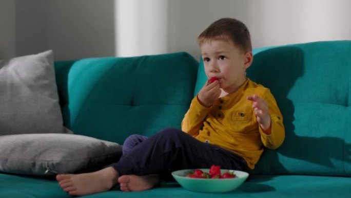 美丽的金发男孩坐在沙发上，在草莓上盛宴