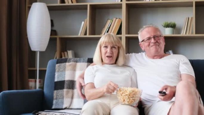 幸福的已婚老人夫妇坐在沙发上看电视，老夫妻吃爆米花笑