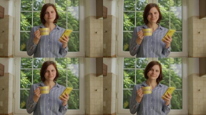 30多岁的女人用手机站在厨房里看着相机，用 “梦” 唱歌喝黄色杯子的咖啡。微笑的女人在家里拿着黄色手