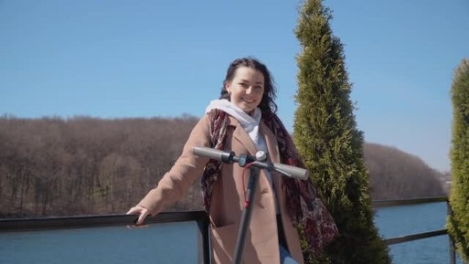 一名年轻的学生女孩带着一辆电动滑板车站在河边的观景台上。女孩呼吸新鲜空气，看着相机。面向年轻人和商人