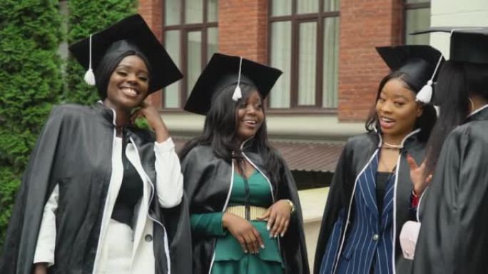 快乐的大学或大学毕业生穿着活泼的斗笠和方形的帽子，生动活泼地交流。非裔美国女学生站在大学大楼附近