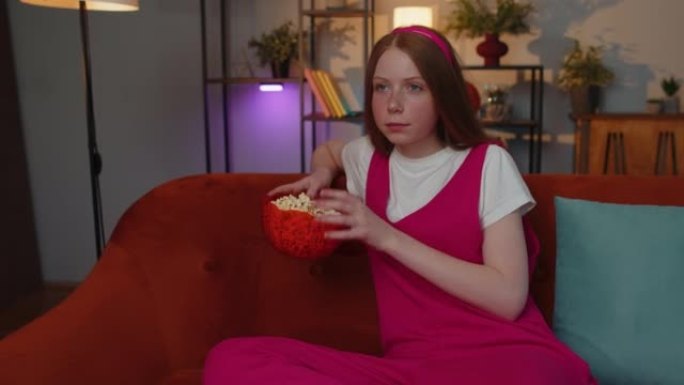 女孩坐在沙发上吃爆米花，在家看有趣的电视连续剧，在线体育游戏