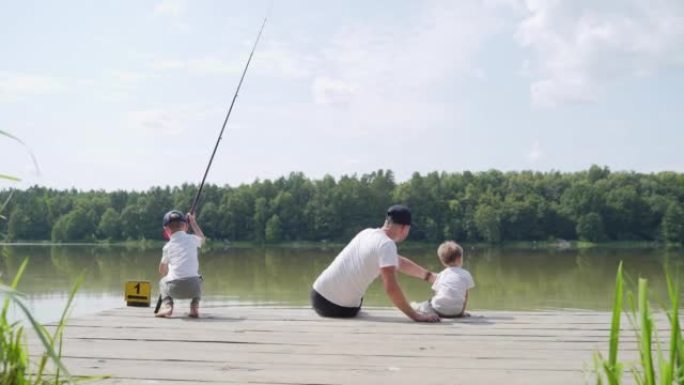 父亲两个小孩儿子抓鱼坐在木桥或码头上。家庭爸爸和孩子钓鱼一起度过休闲时间露营教钓鱼