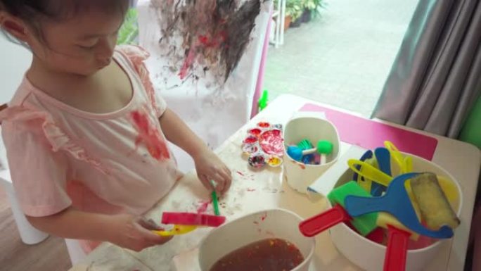 亚洲女童站在游戏室的画架上的白色画布上绘画。尝试在桌子上使用绘图工具。儿童的创造力。家里画过程这么脏